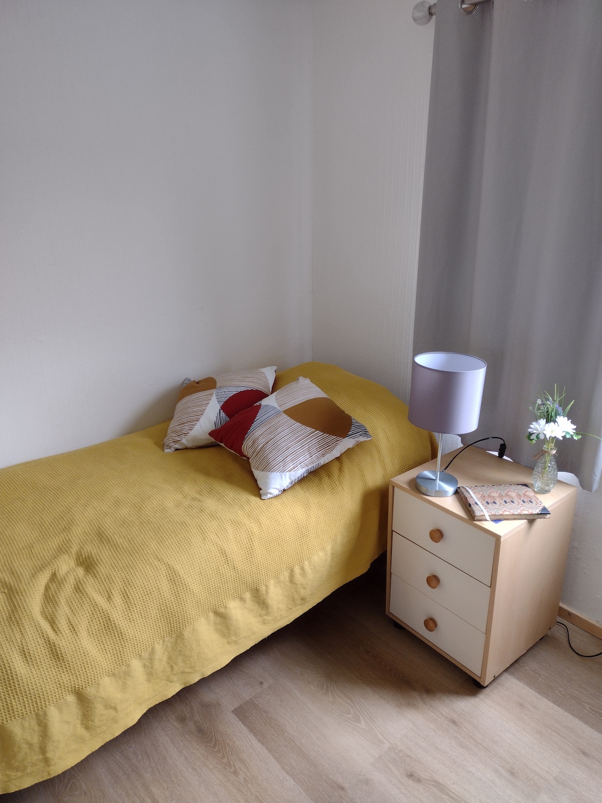 A single bed is covered with a yellow knitted blanket, accented with patterned cushions. A bedside table, featuring three drawers, holds a decorative lamp and a small vase with flowers. Natural light enters through a nearby window adorned with light gray curtains.