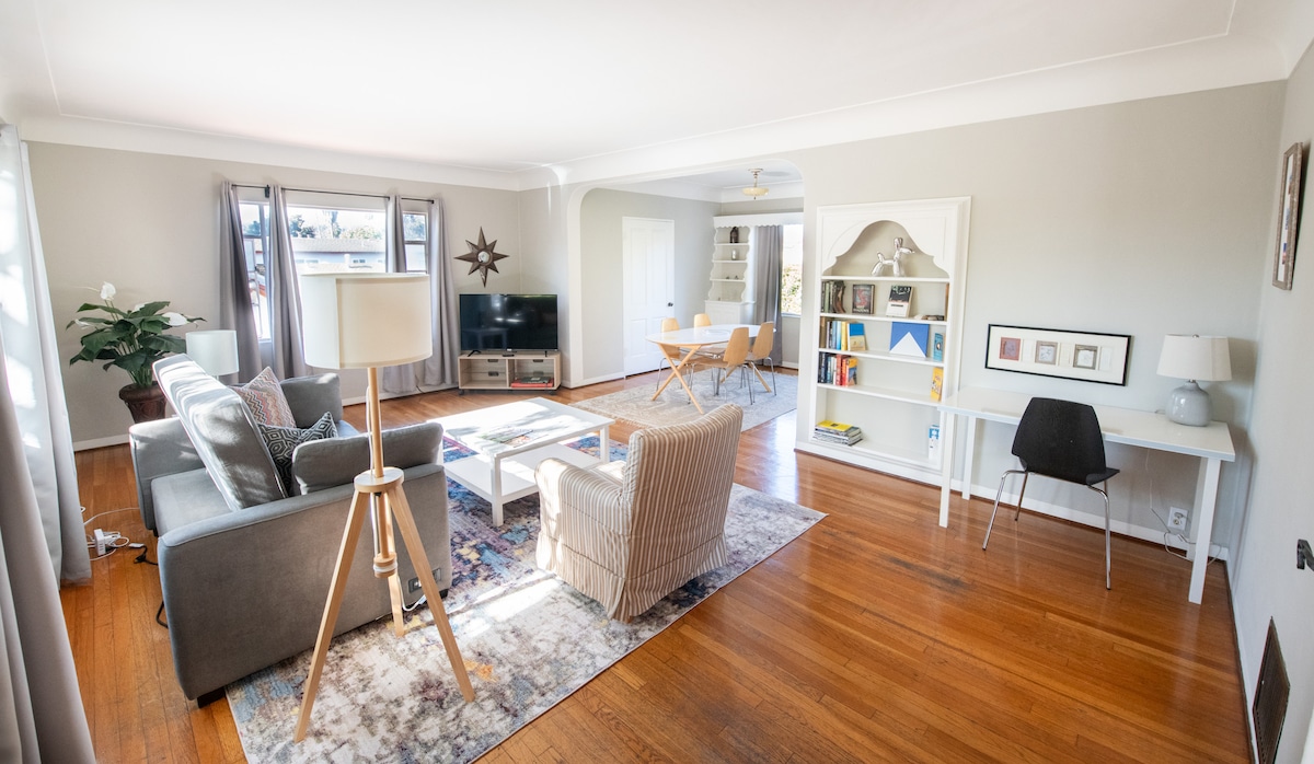The spacious living area features a combination of comfortable seating, including a sectional sofa and an armchair. A coffee table sits on a light area rug, while a large window allows natural light to brighten the room. A desk and chair are present by a bookcase.