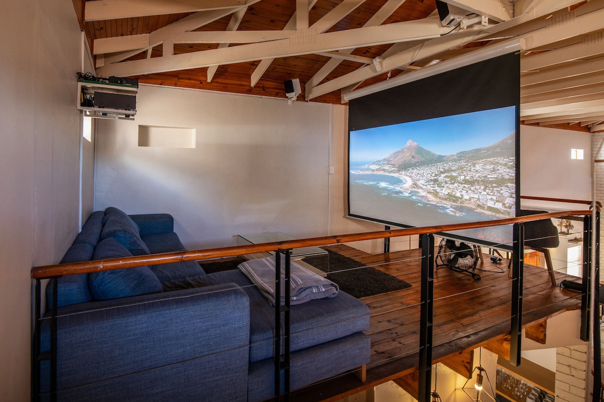 A spacious mezzanine area features an L-shaped couch and a large screen displaying a scenic coastal view. The wooden floor adds warmth, while exposed beams highlight the high ceiling, creating a comfortable space for relaxation and entertainment.