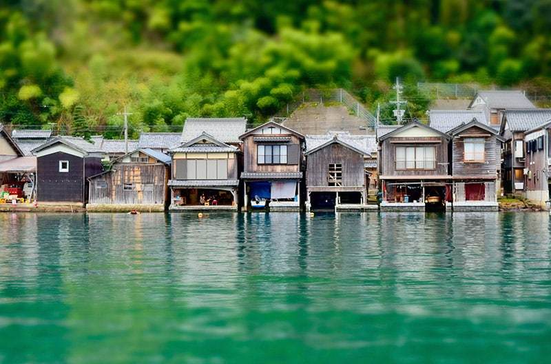 A row of traditional wooden boathouses lines the serene water, reflecting the lush greenery in the background. Each structure features a unique design, with some having open doors revealing boats secured within. The calm water enhances the peaceful setting.