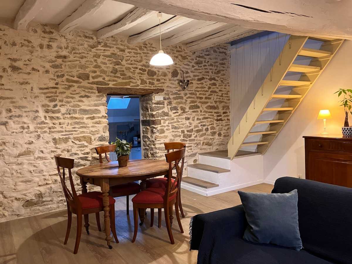 An inviting living space features a round wooden dining table surrounded by four chairs, set against a backdrop of exposed stone walls. A spacious staircase leads to the upper level, while soft lighting is provided by a pendant lamp.