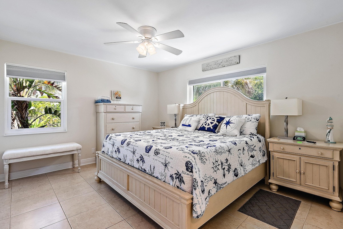 A spacious bedroom features a large bed adorned with a coastal-themed quilt. Two windows allow natural light to brighten the room, framed by simple blinds. A dresser and bedside tables provide ample storage, complementing the light decor and tiled flooring.