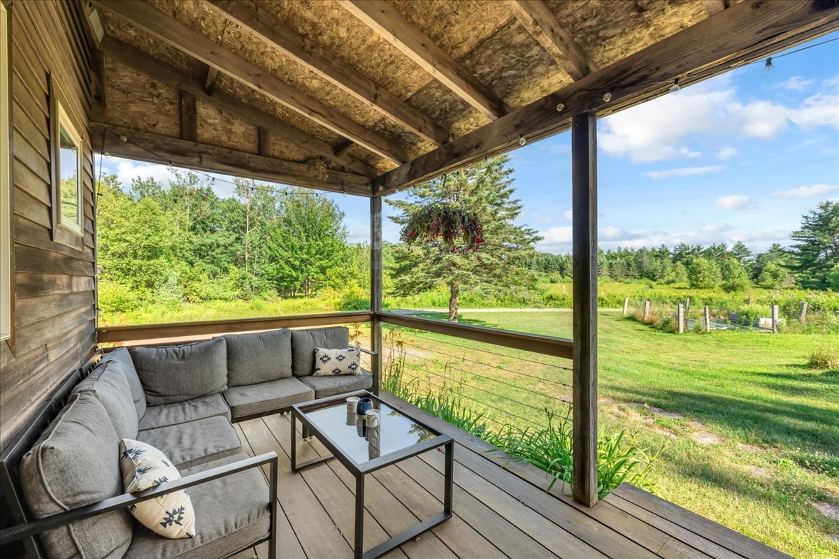 A covered porch features a comfortable seating area, with a sectional couch and a glass coffee table. Lush greenery surrounds the area, with views of the expansive field and trees in the background. Natural light floods the space, creating a serene atmosphere.