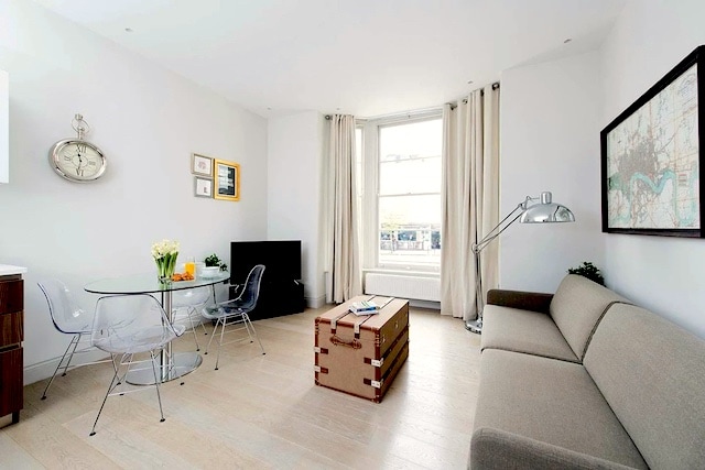 A bright and airy living area features large windows with sheer curtains, allowing natural light to enter. A modern glass dining table is paired with four transparent chairs. A plush sofa and a decorative trunk serve as functional furniture, while a wall-mounted TV completes the space.