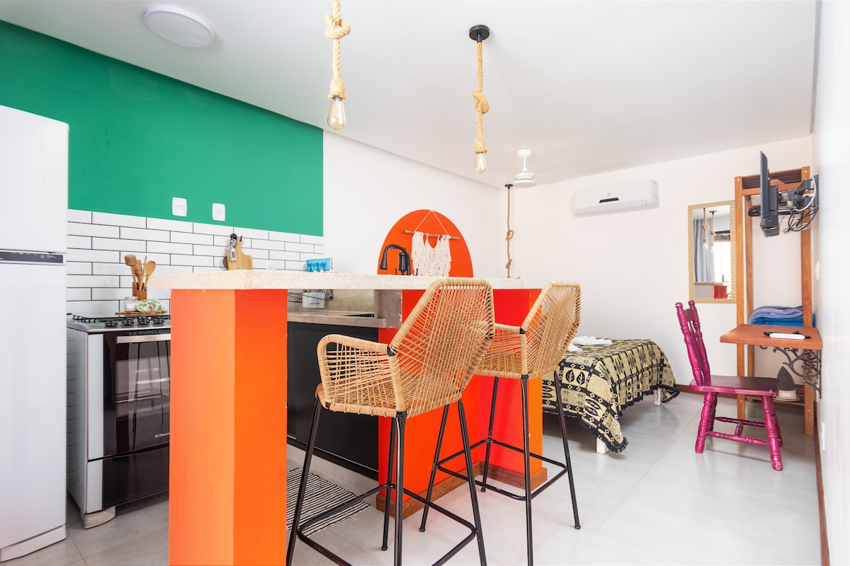 A modern kitchenette features a vibrant green accent wall and a sleek bar with three woven stools. An adjoining bed area is visible, with a colorful blanket draped over the bed. Natural light brightens the space through a nearby window, enhancing the open layout.