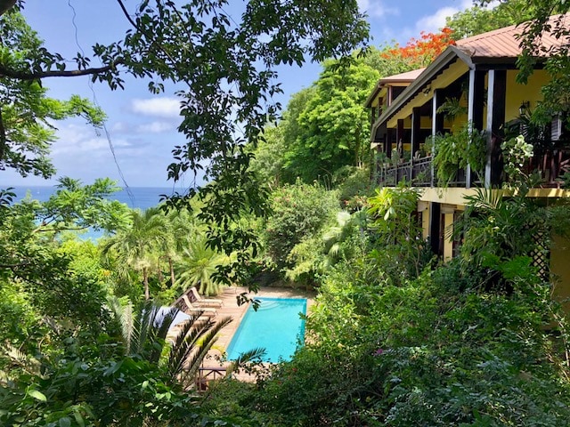 Surrounded by lush greenery, a view of the inviting swimming pool is visible at the center, framed by tropical plants. The multi-level villa overlooks the pool area, with the Caribbean ocean seen in the distance beneath a clear sky.
