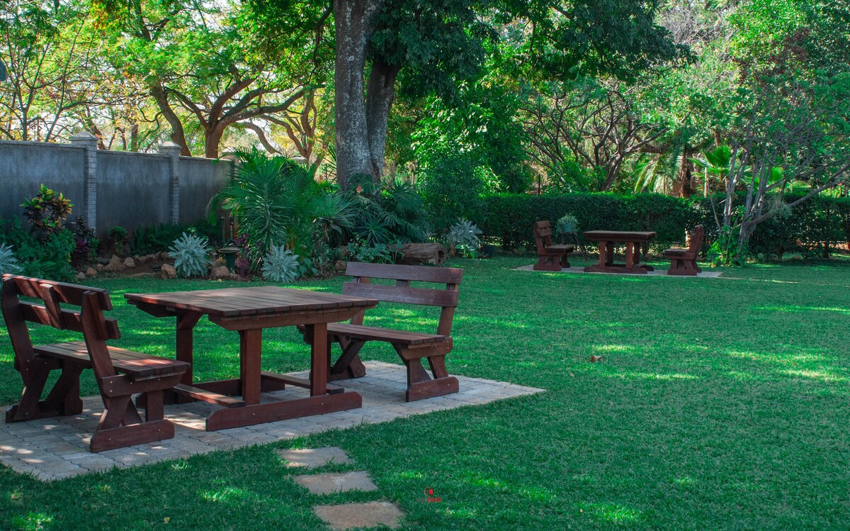 A shared garden area features well-maintained grass, with wooden picnic tables and benches arranged under the shade of trees. Lush greenery surrounds the seating areas, providing a peaceful outdoor space for guests to enjoy.