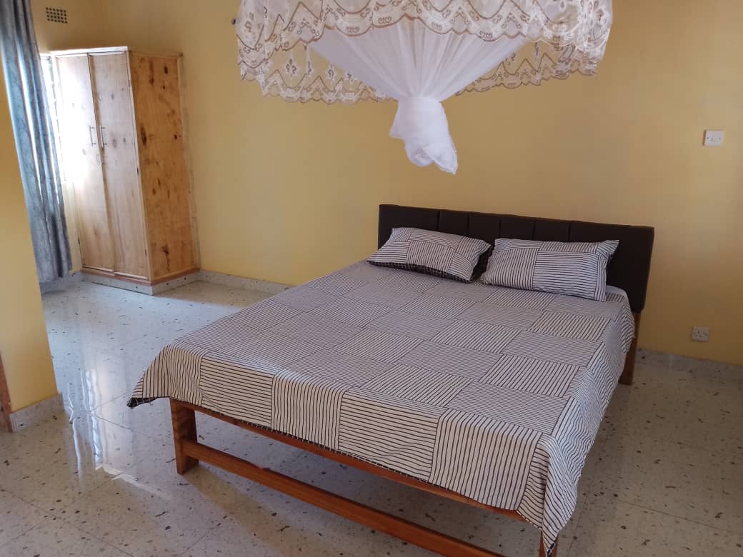 A spacious bed with a striped blanket is positioned in the center of the room, accompanied by two pillows. A wooden wardrobe is visible in the background, while soft light filters through a window adorned with light curtains.