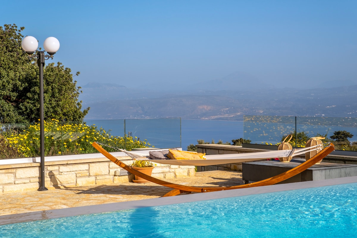 A serene outdoor space features a wooden hammock set beside a private swimming pool. The scene includes clear views of the sea and distant mountains under a bright blue sky. Yellow flowers and greenery surround the area, enhancing the tranquil atmosphere.