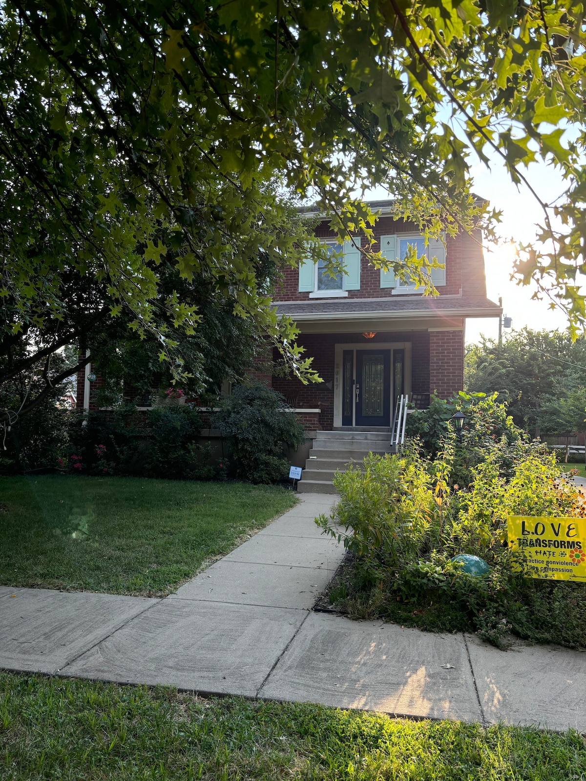 A two-story brick building is framed by leafy trees and well-maintained greenery. A concrete pathway leads to the entrance, which features steps and a small landing. A vibrant garden sign is visible, adding a touch of character to the inviting exterior.