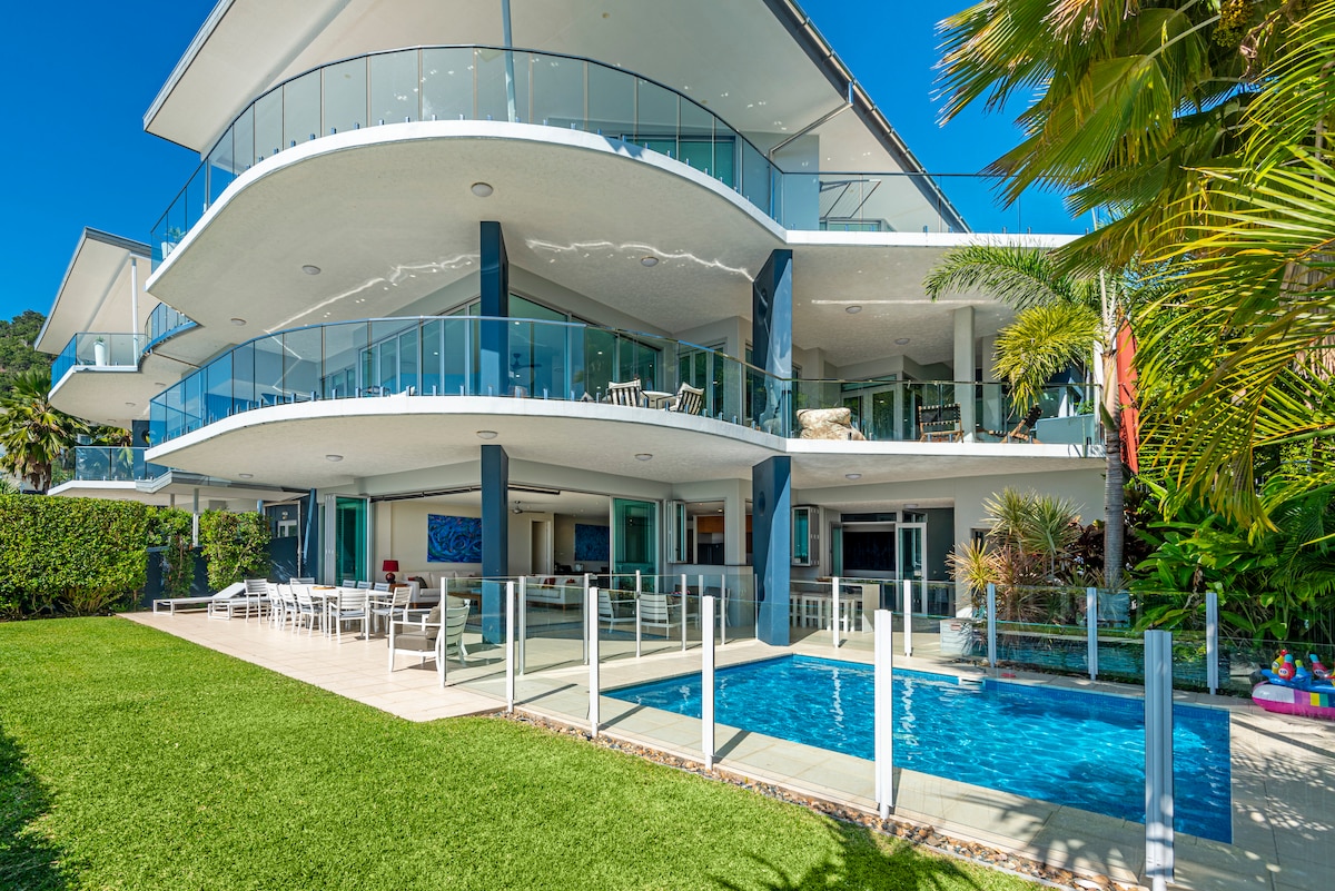 A modern multi-story building features a large outdoor entertaining area with ample seating. An in-ground swimming pool is surrounded by glass fencing, and lush greenery frames the space, enhancing the connection to the outdoor environment.
