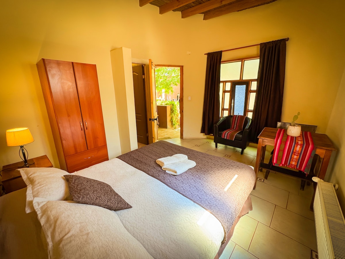 A well-lit room features a spacious bed dressed in soft linens, complemented by a cozy armchair and a wooden desk. A tall wardrobe stands against the wall, enhancing the room's functionality. Natural light enters through large windows adorned with dark curtains.