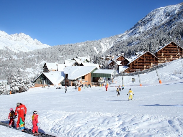 A snowy landscape is depicted, with several ski slopes and a variety of skiers, including children, navigating the terrain. Surrounding chalets are visible, set against a backdrop of densely forested mountains. The clear blue sky adds to the crisp winter ambiance.