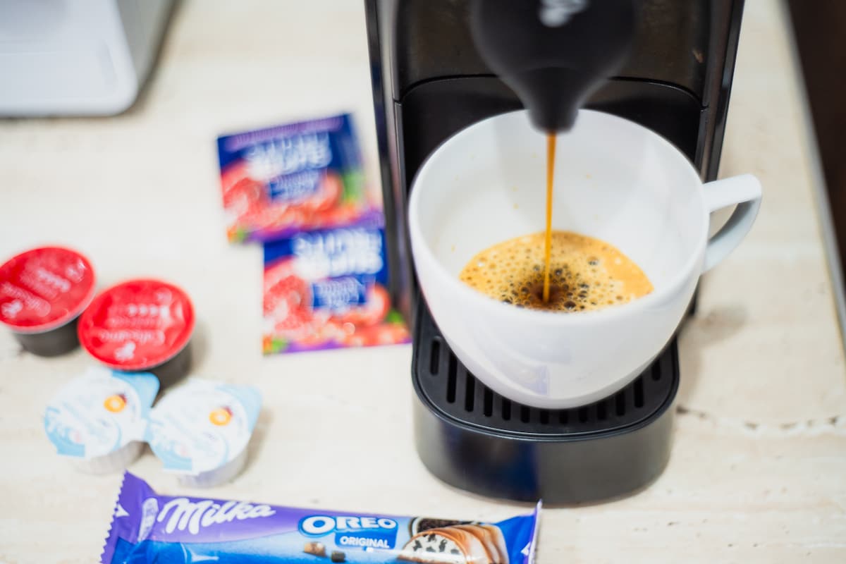 A coffee maker is seen preparing a fresh cup of coffee into a white mug. Nearby, various coffee condiments and treats are displayed, including two coffee creamers, sugar packets, and chocolate snacks. The countertop offers a functional and inviting setting for morning routines.