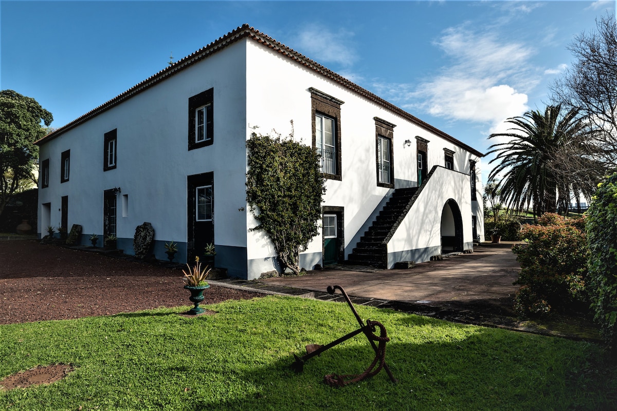 The exterior of a white 17th-century manor house is shown, featuring dark green window shutters and a staircase leading to the upper level. The property is surrounded by well-maintained gardens, with palm trees visible in the background and a decorative anchor placed on the grass.