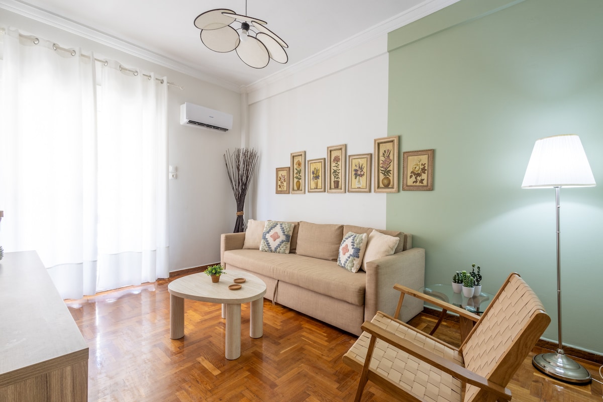 A light-filled living area features a beige sofa adorned with patterned cushions, complemented by a wooden coffee table. Framed botanical art pieces line the wall, adding a touch of nature. A modern floor lamp stands near a wooden armchair, enhancing the space's inviting ambiance.