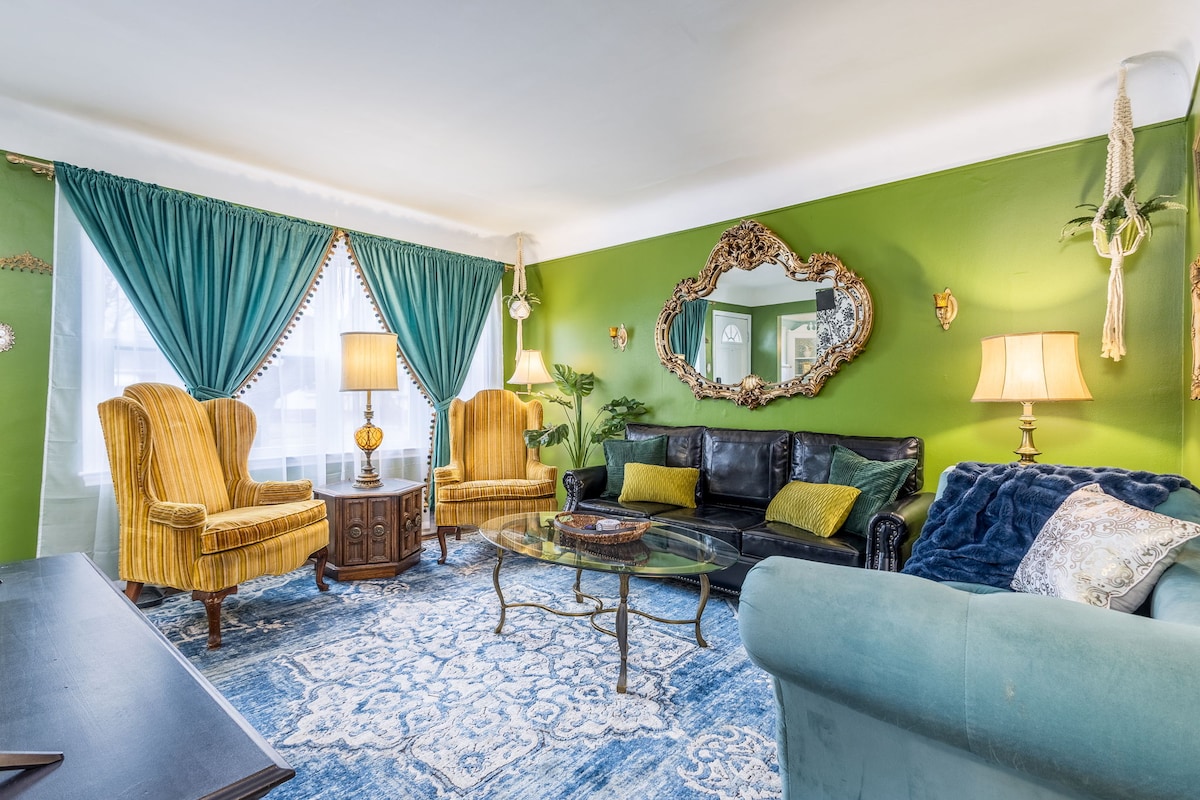 The living room features a vibrant green wall, complemented by elegant blue drapes. A black faux leather couch is paired with a glass coffee table and vintage accent chairs in yellow. Soft lighting is provided by table lamps, adding to the cozy ambiance.