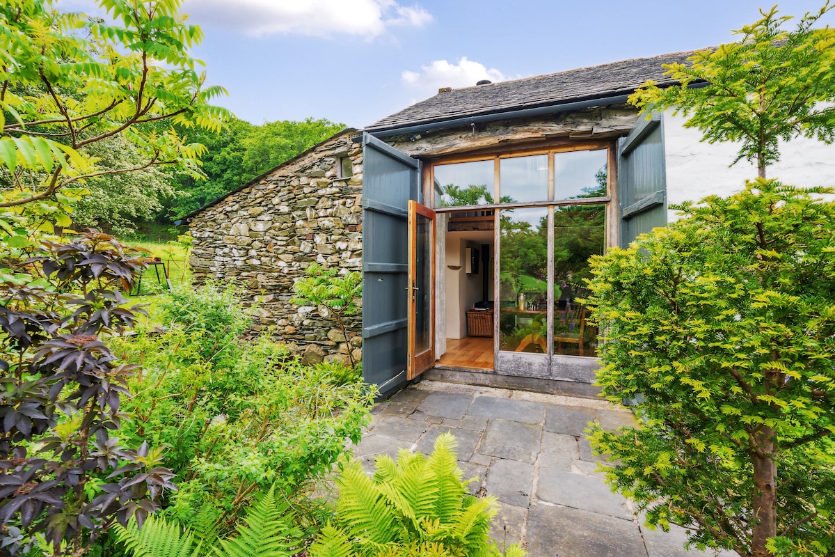 A charming entrance is framed by expansive glass doors, opening onto a patio area. Lush greenery surrounds the entrance, with various plants and trees enhancing the natural setting. Stone walls complement the rustic aesthetic of the cottage, blending seamlessly with the landscape.