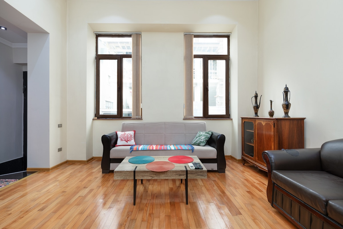 The living room features two sofas, a coffee table adorned with colored circles, and a wooden dresser. Natural light enters through multiple windows, highlighting the polished wooden floor and high ceilings.