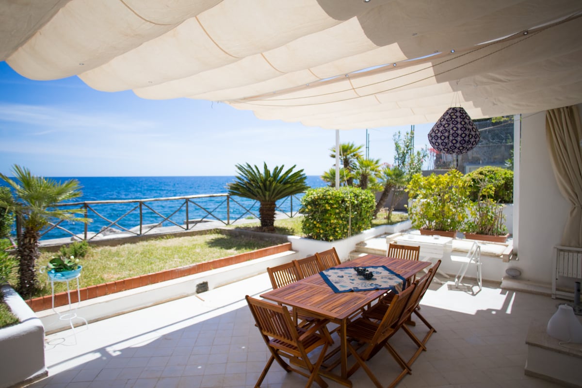 A spacious terrace overlooks the sea, featuring a wooden dining table surrounded by several chairs. Lush greenery and palm trees frame the area, while a fabric canopy provides shade. A decorative pendant light hangs above, enhancing the outdoor dining experience.