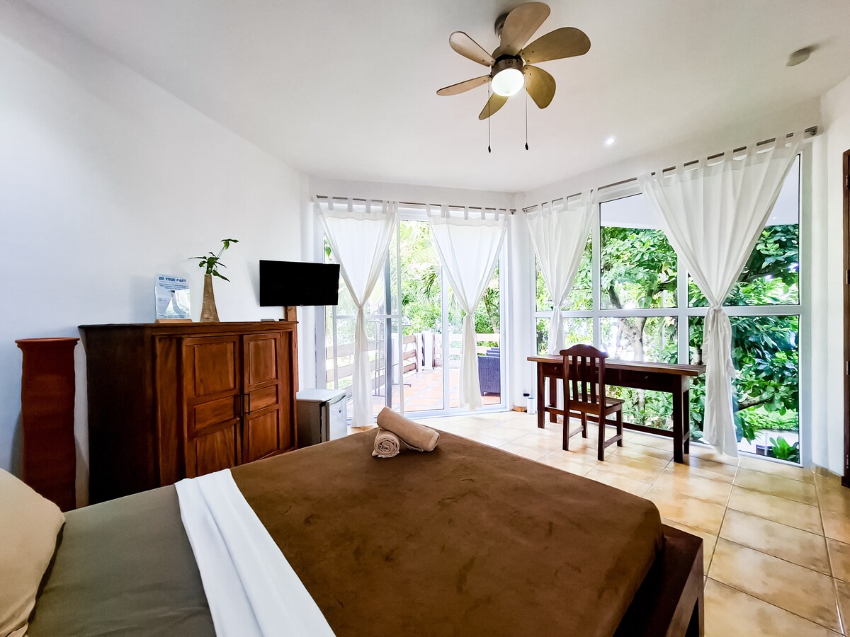 A spacious room features large glass doors that open to a tranquil garden view. Natural light fills the area, accentuating the warm tones of the furniture, including a wooden chest and a small desk. A comfortable bed is neatly arranged with soft linens.