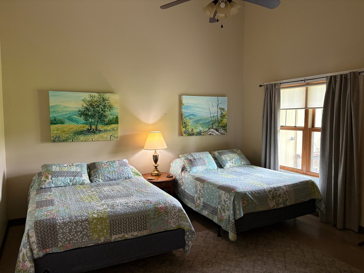 The bedroom features two queen beds, each with colorful patchwork bedding, complemented by wooden nightstands. Two landscape paintings adorn the walls, and a lamp provides gentle illumination. Natural light filters through a large window, framed by light gray curtains, enhancing the room's spacious feel.