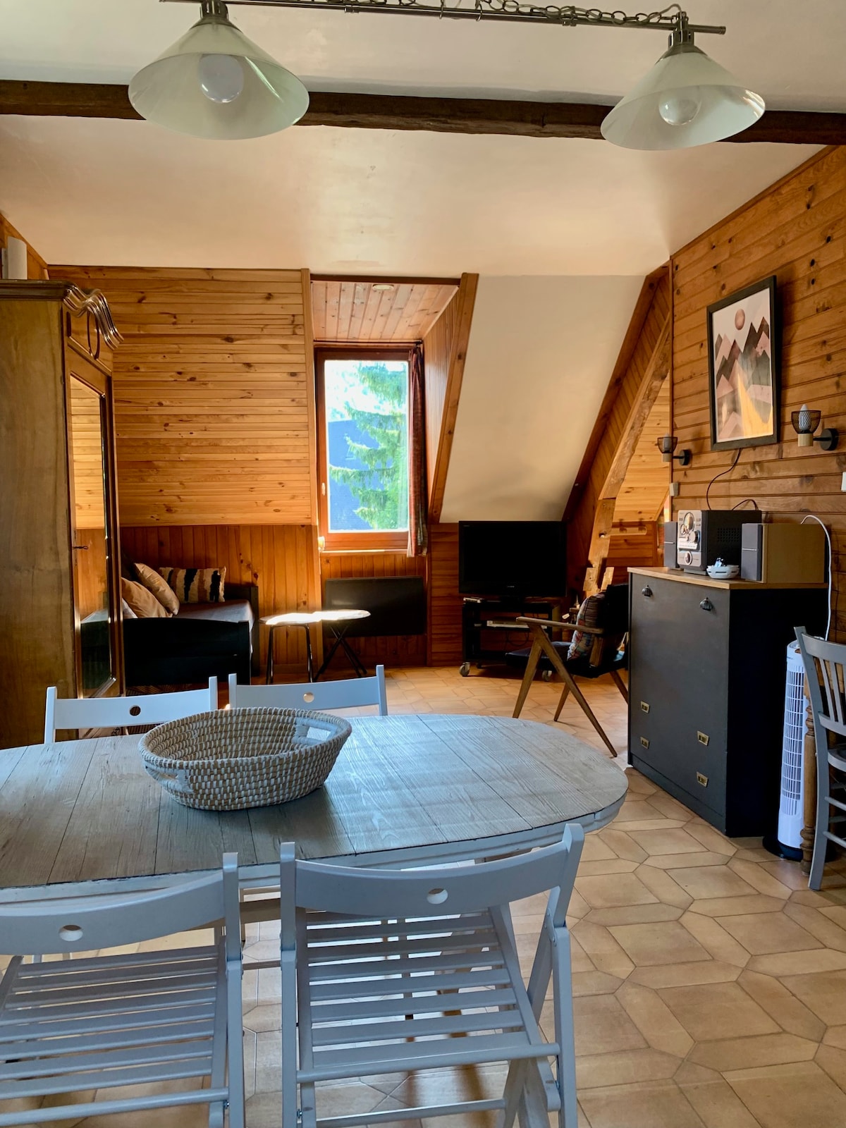 A cozy living space features wooden walls and angled ceilings, creating a warm atmosphere. A dining table with four chairs is positioned at the center. A small coffee table and a sofa are placed nearby, with a television on a cabinet across from them. Natural light enters through a window.