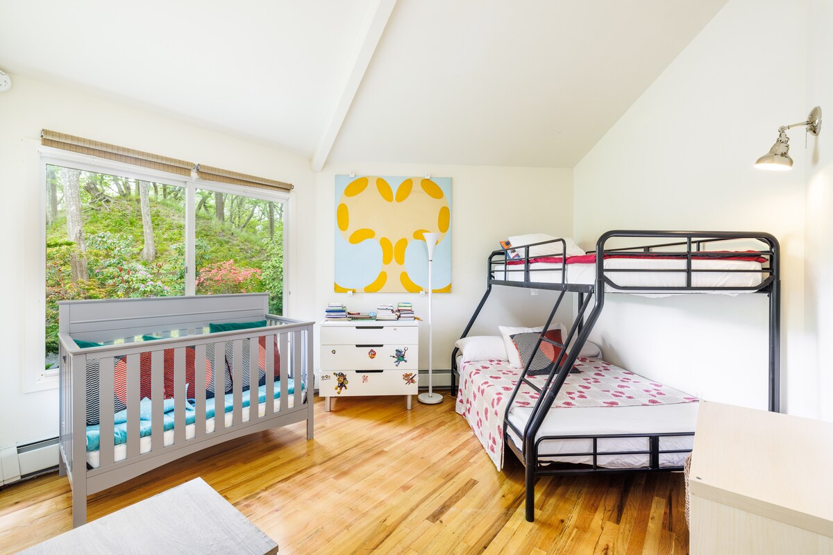 A well-lit bedroom showcases a combination of a crib and a bunk bed, providing options for various sleeping arrangements. A single bed is also present, with colorful bedding adding a playful touch. A large window invites natural light and offers views of the surrounding greenery.