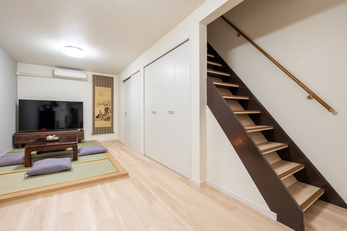 A living area features a traditional low table surrounded by floor cushions on tatami mats. A large television is mounted on the wall, and a sliding door leads to a storage area. Stairs to the upper level are visible, along with a bright overhead light.