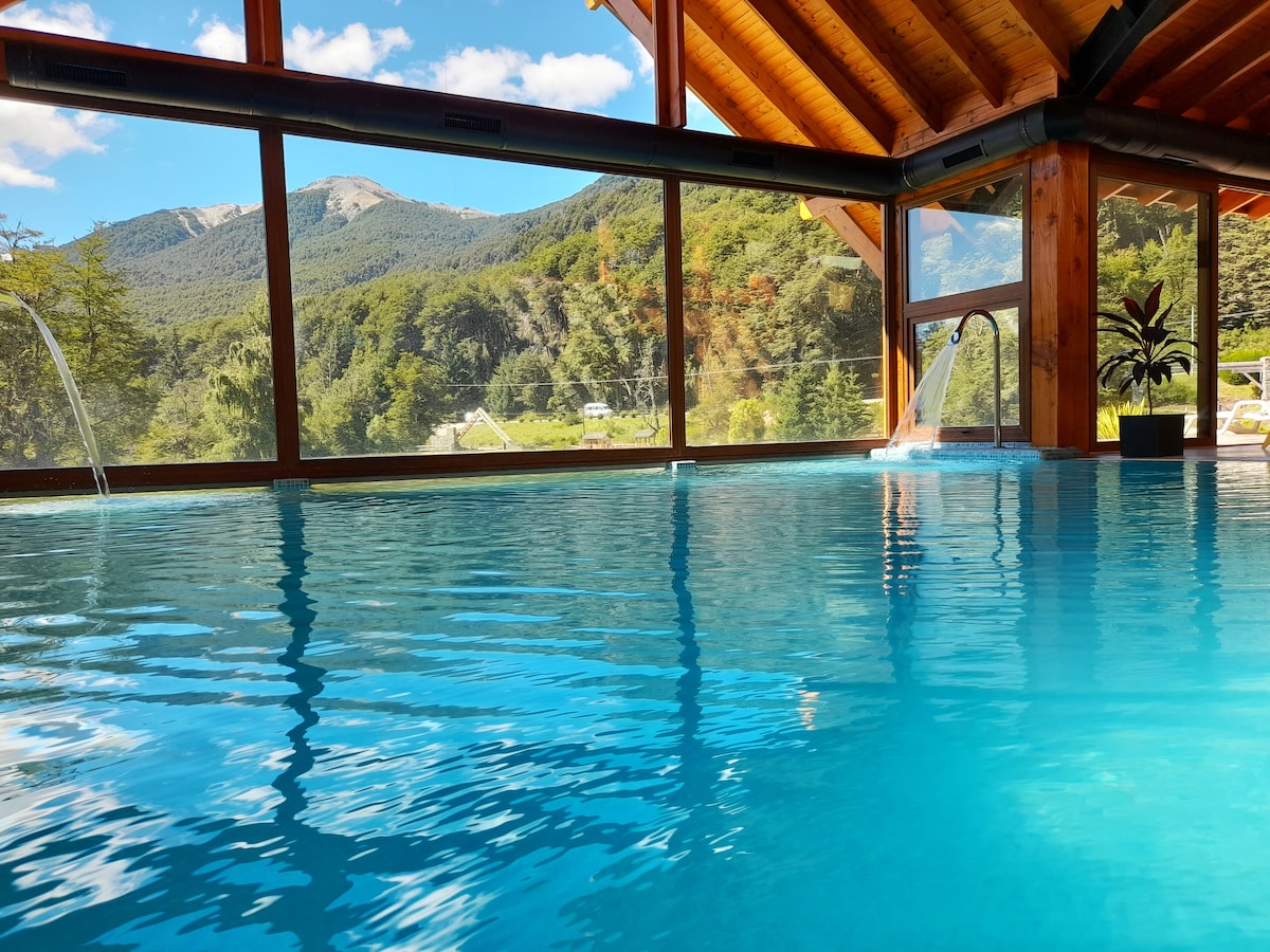 An indoor pool is seen with clear, turquoise water, surrounded by large windows that offer a view of the mountains and greenery outside. Natural light illuminates the wooden beams of the ceiling, enhancing the serene setting.