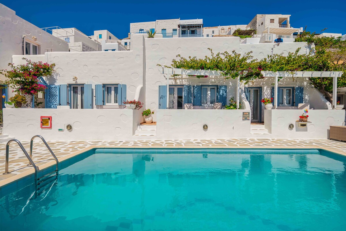 A bright exterior showcases a traditional white building adorned with blue window shutters and vibrant floral accents. A clear swimming pool reflects the blue sky, while steps lead into the water, creating an inviting atmosphere for relaxation.