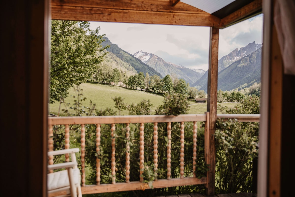A view from the balcony offers a sweeping landscape of lush green fields framed by trees, with distant mountains displaying patches of snow. The natural scenery provides a calming sense of space and connection to nature.