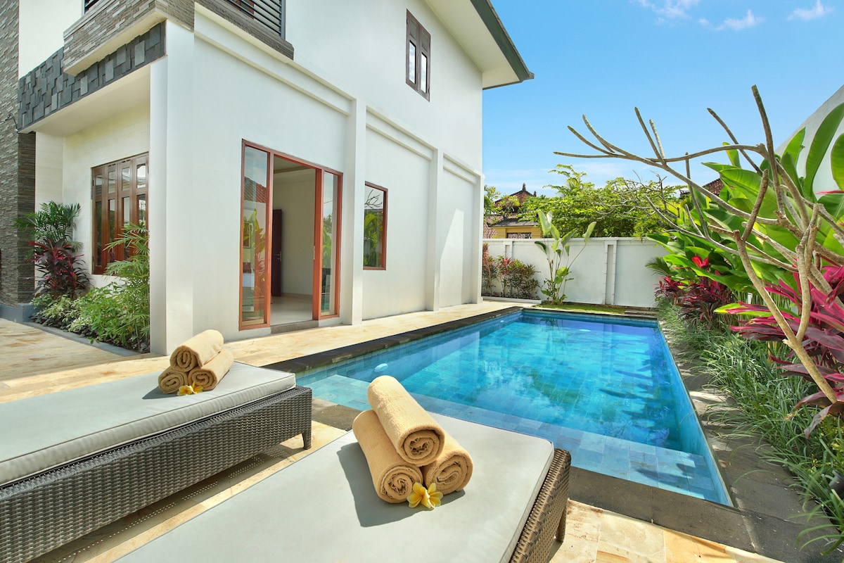 A serene outdoor area features a crystal-clear pool surrounded by tropical greenery. Two lounge chairs with rolled towels rest by the poolside, inviting relaxation under the sun. Bright blue skies and delicate plant foliage enhance the tranquil ambiance.
