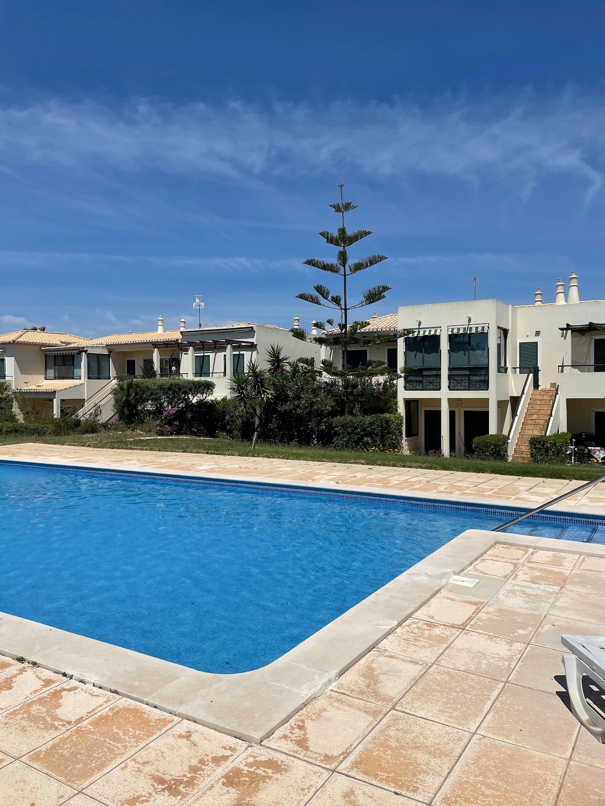 A swimming pool, reflecting the clear blue sky, is framed by stone tiles and bordered by well-maintained gardens. The apartment complex features multiple units with green shutters and balconies, creating a serene atmosphere for relaxation. Lush trees provide shade along the poolside.