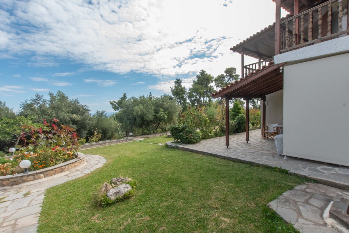A spacious outdoor area features a stone path leading through lush greenery and colorful flower beds. A stone terrace provides access to the cottage, with a shaded seating area visible under the balcony above. The sky is lightly clouded, enhancing the serene environment.