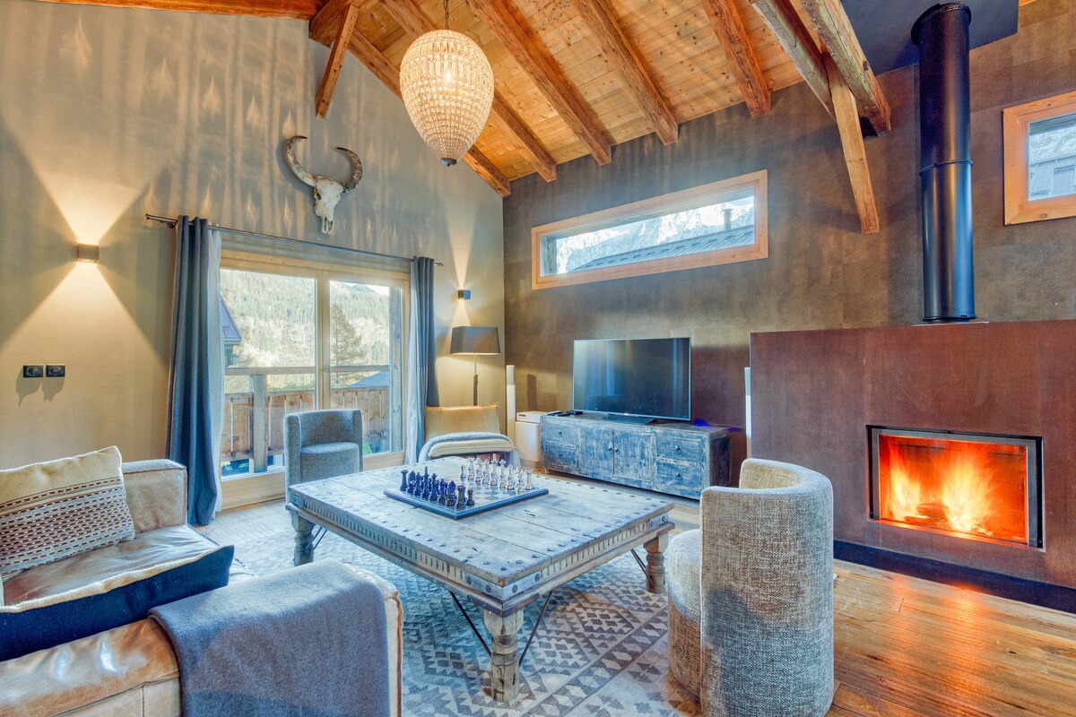A spacious living room features high wooden beams and a warm fireplace, offering a cozy ambiance. A large, patterned area rug sits under a coffee table with a chess set. Natural light filters through wide glass doors that open to mountain views.
