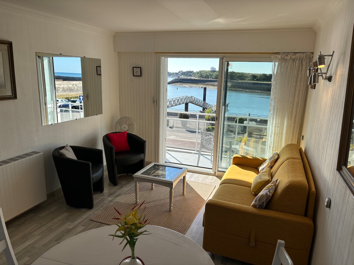 A bright living area features large glass doors opening to a view of the channel. Comfortable seating includes two black chairs and a yellow sofa. A glass coffee table sits on a neutral rug, while a small fan is positioned nearby. Natural light enhances the space.