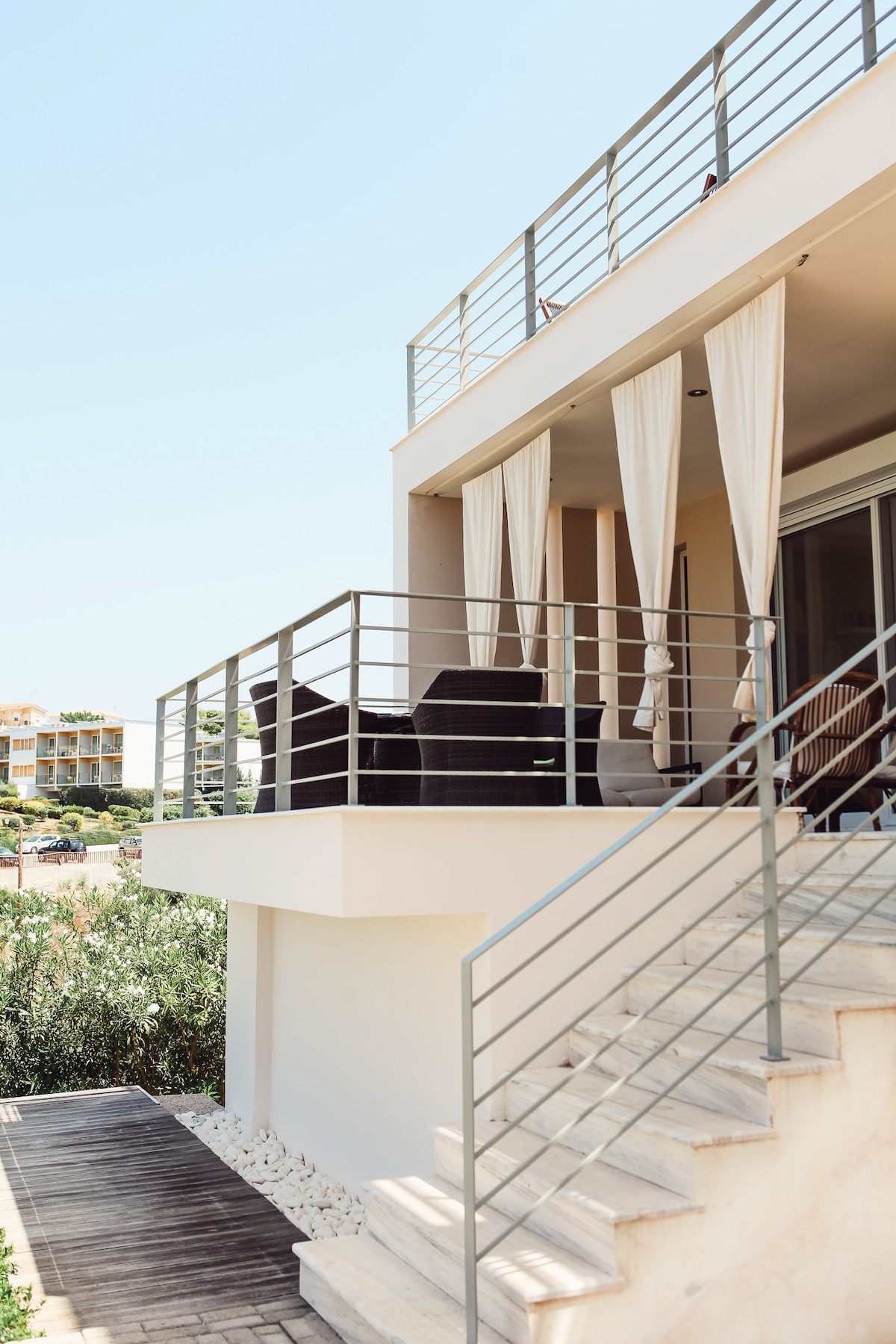 An outdoor terrace area is visible, featuring a railing and draped curtains for privacy. Seating arrangements include dark wicker chairs, positioned to enjoy the sunny ambiance. Stairs lead down to a well-kept pathway lined with stones, enhancing the connection between indoor and outdoor spaces.