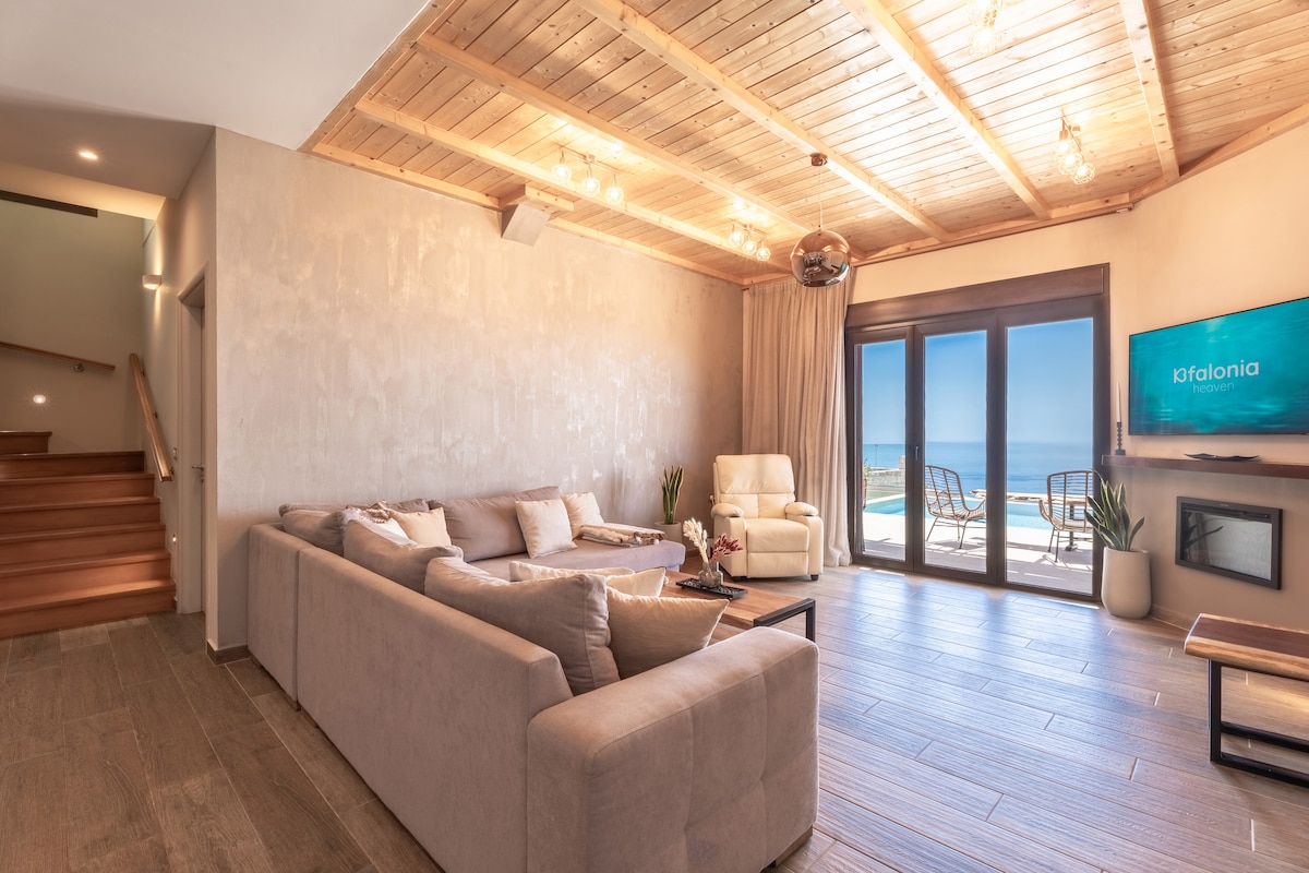 A spacious living area is featured, highlighting a large, comfortable sectional sofa and two armchairs facing expansive glass doors that open to a view of the outdoor terrace and sea. Warm wooden beams span the ceiling, and a television is mounted on the wall.