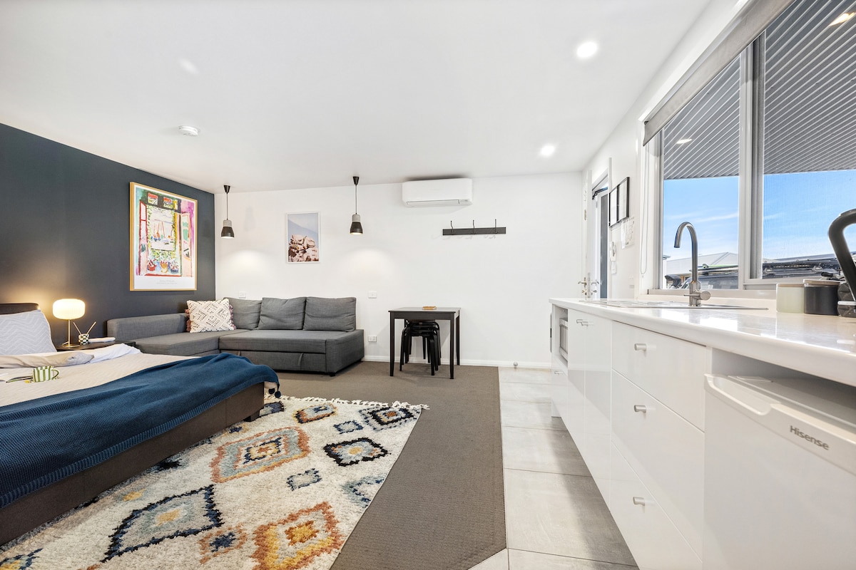 A spacious studio apartment features a cozy seating area with a large gray sofa, a dining table, and a kitchenette. Light streams in from a large window, illuminating the contemporary decor and patterned area rug, creating a functional and inviting living space.