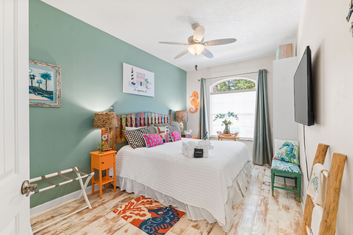 A serene and colorful bedroom with a Seahorse theme, featuring a beautifully crafted handmade and hand painted Shoestring Creations king-sized headboard, Mackenzie Childs pillows, and ocean-inspired decor. 