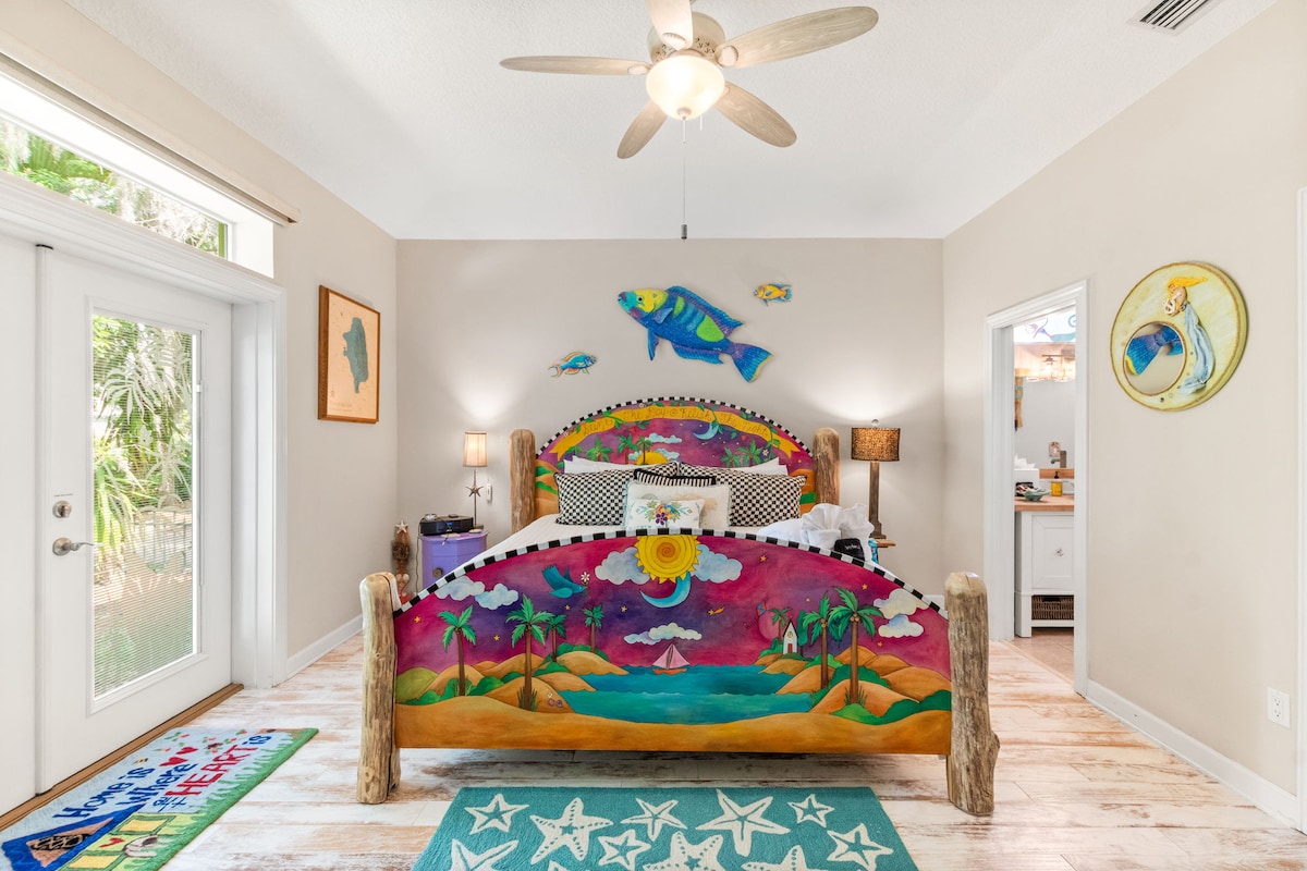 This vibrant and artistic bedroom is a coastal retreat, featuring a beautifully hand-painted bed by Sticks, with tropical scenes that evoke the serenity of a seaside escape. The room is adorned with colorful coastal-themed art.