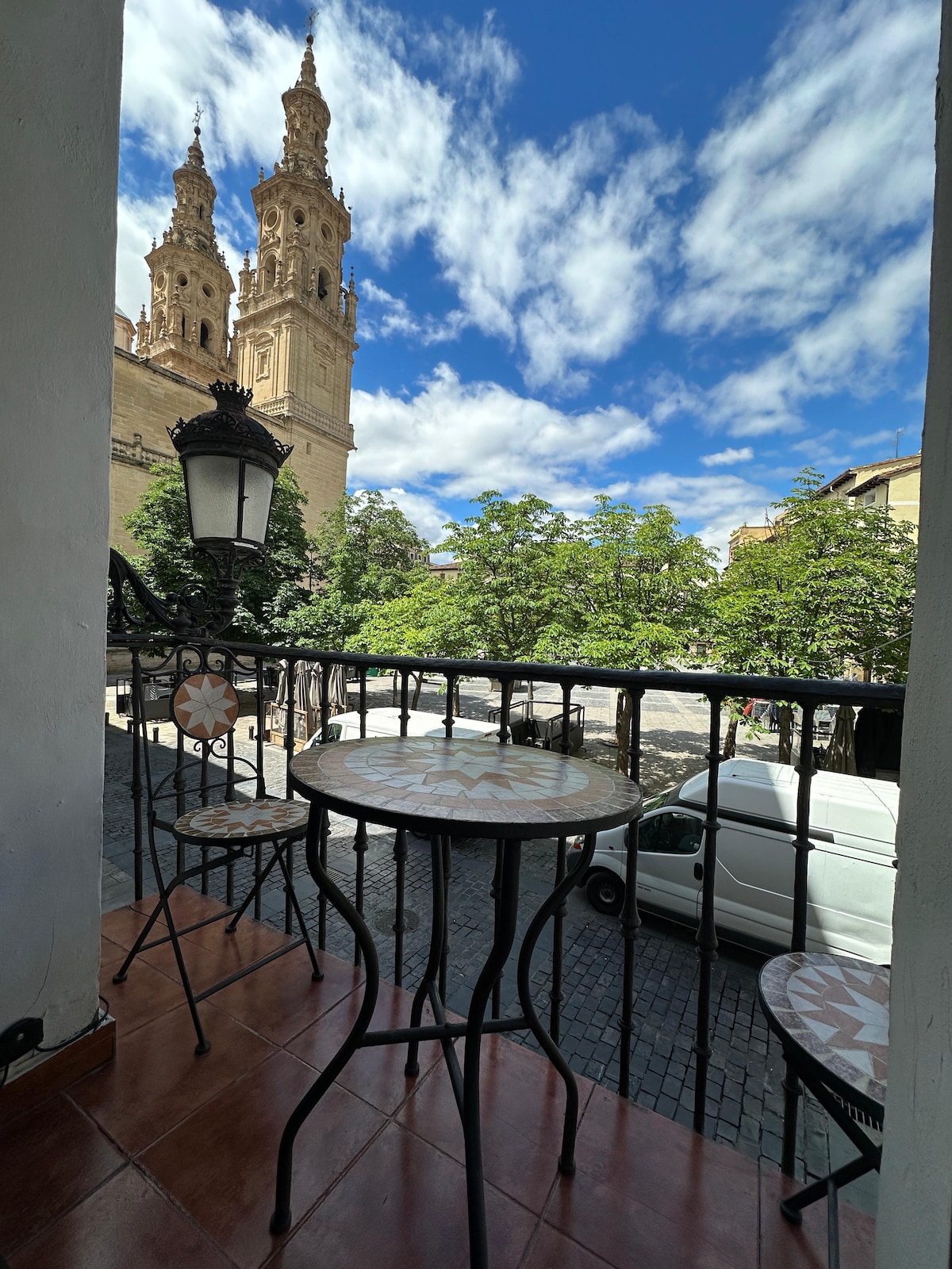 A small balcony offers a table and two chairs with decorative mosaic patterns. Views of a stunning cathedral tower are framed by vibrant greenery and a partly cloudy blue sky, creating an inviting outdoor space perfect for relaxation.