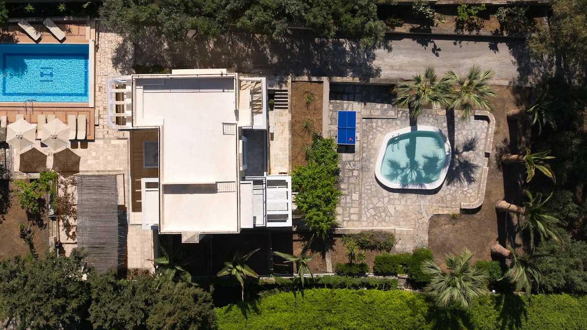 An aerial view of the OLIVETUM villa highlights two swimming pools surrounded by landscaped gardens. The larger pool is positioned beside sun loungers, while the shallower pool is nestled within a stone patio area. Lush greenery and palm trees frame the outdoor space, creating a serene atmosphere.