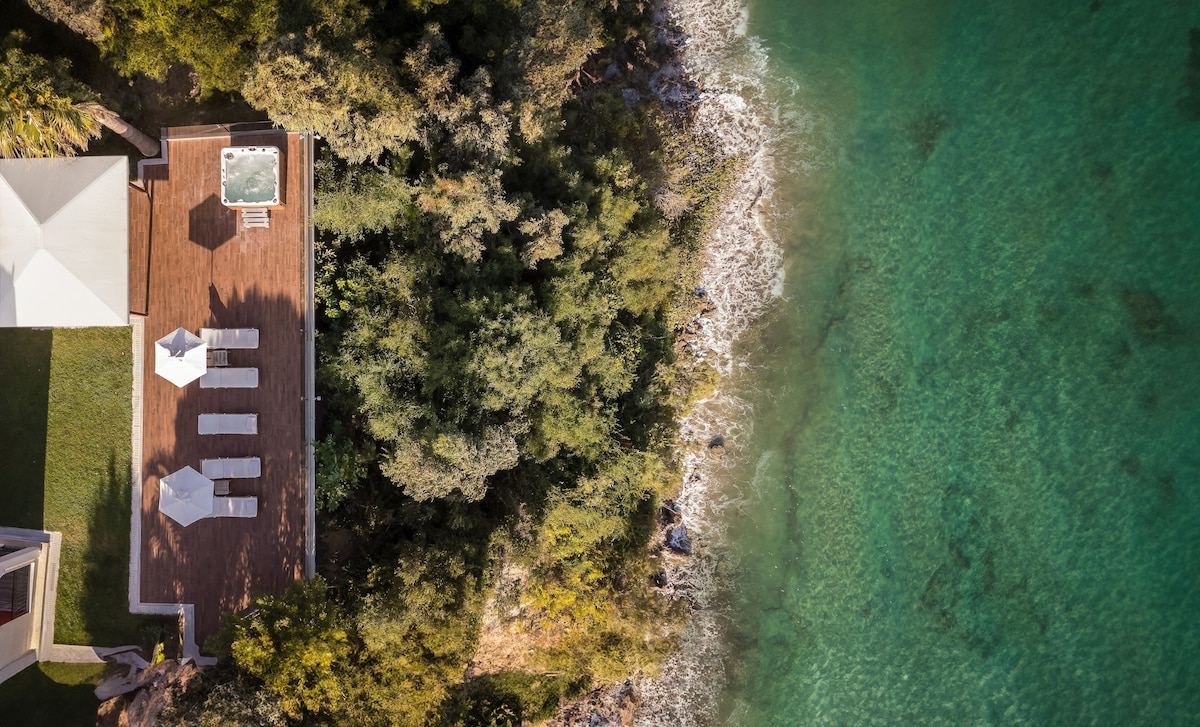 An aerial view showcases a wooden deck alongside a clear green-blue sea, with sun loungers positioned under white umbrellas. Lush greenery borders the area, providing a natural contrast to the shoreline where soft waves gently lap against the rocks.