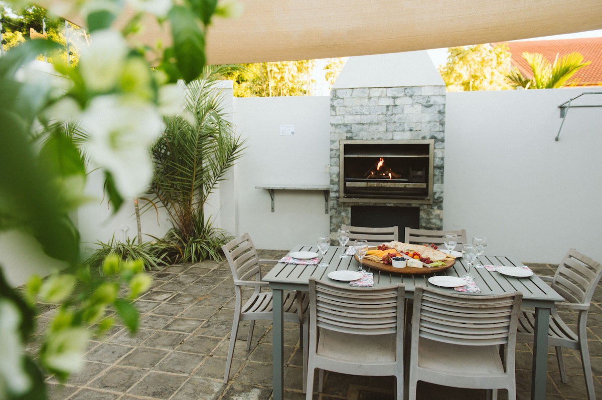 An outdoor dining area is presented with a rectangular table surrounded by light-colored chairs. A barbecue grill is positioned against a stone wall, with a fire gently burning inside. Green foliage frames the space, adding a touch of nature.