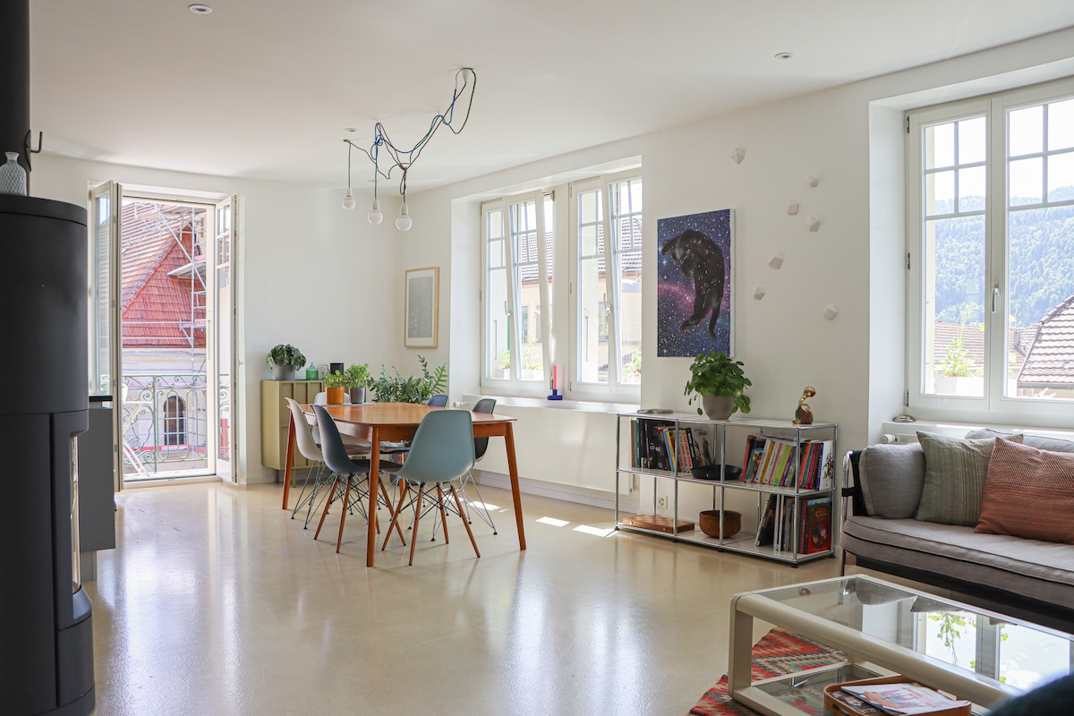 A bright living area features large windows opening to a balcony, providing ample natural light. A dining table with chairs is positioned centrally, while a comfortable sofa is arranged nearby. Various plants are placed throughout the space, contributing to the inviting ambiance.