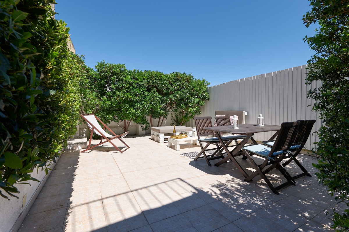 A spacious terrace features various seating options, including loungers and a dining table. Surrounding greenery provides privacy, while a bright blue sky creates an inviting outdoor atmosphere. The tiled floor enhances the open feel of the space, suitable for relaxing or dining al fresco.