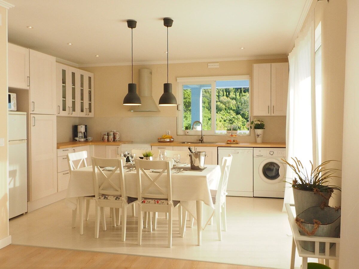 A modern kitchen features white cabinetry and a spacious dining area with a large table surrounded by white chairs. Dual pendant lights hang above the table. Large windows bring in natural light and provide a view of the surrounding greenery.