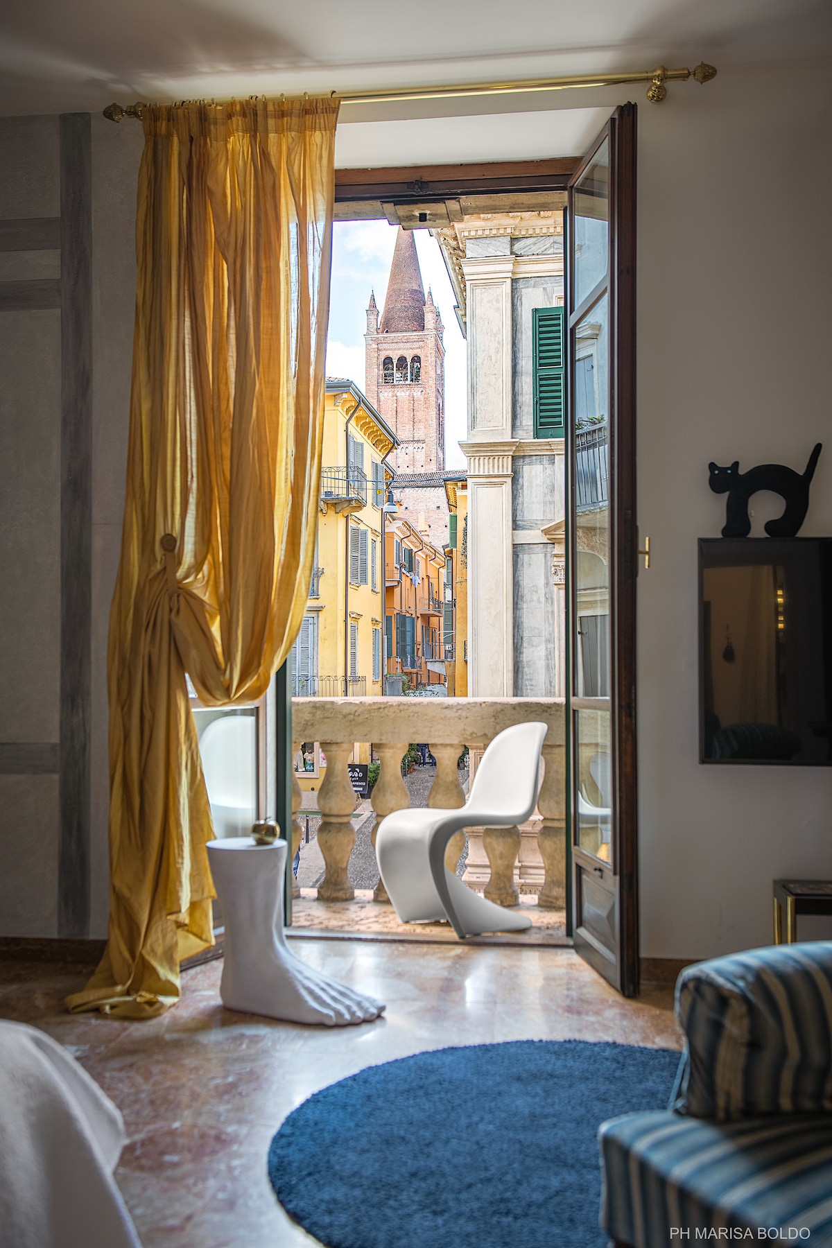 A view from the apartment's balcony reveals a lively street scene in Verona, framed by flowing golden curtains. A modern white chair is positioned near the open door, with a glimpse of the city's distinctive bell tower in the background.